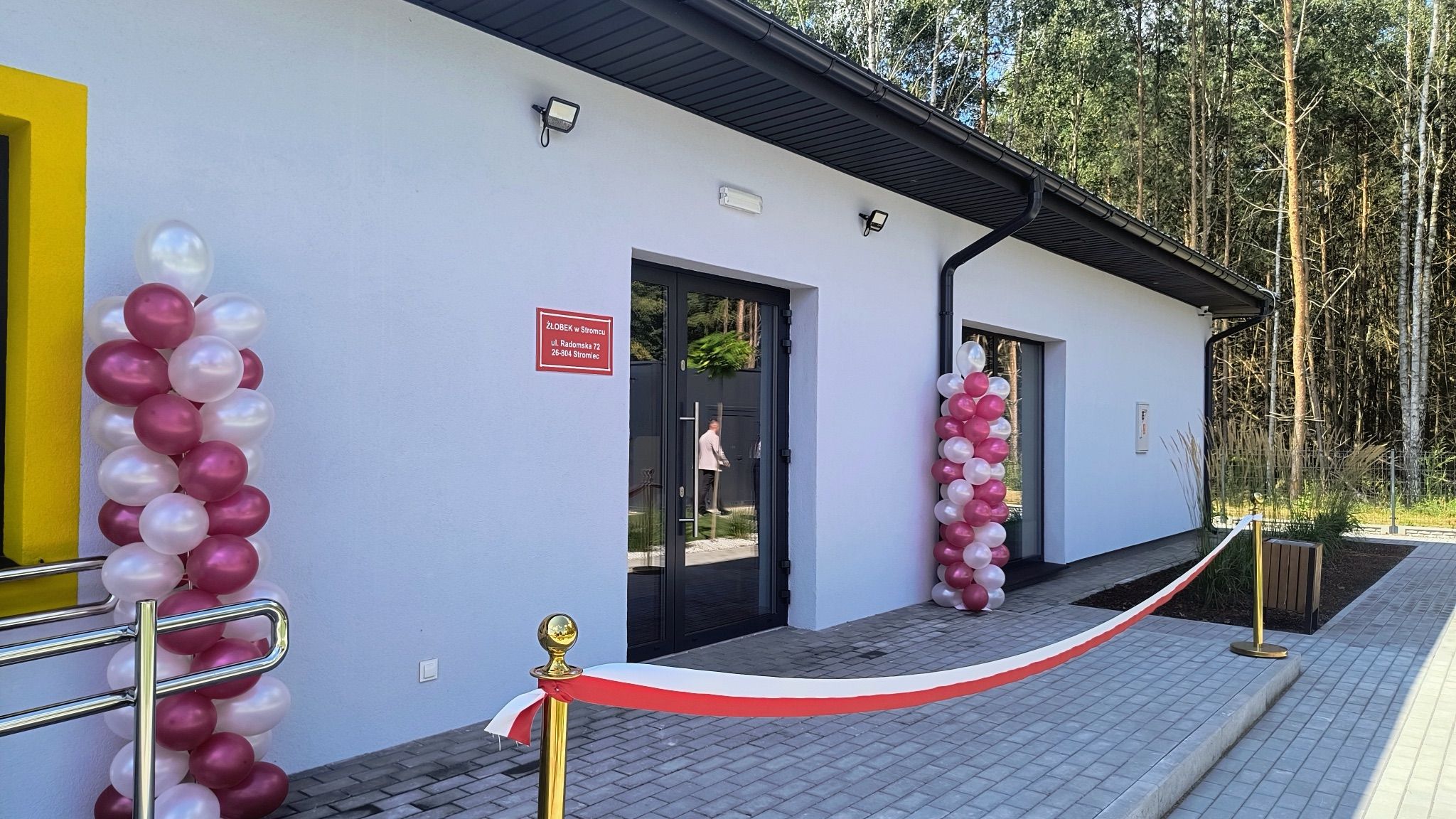 Wejście do żłobka przewiązane biało-czerwoną wstęgą, po bokach kolumny z białych i bordowych balonów, na ścianie tablica z napisem Żłobek w Stromcu, ulica Radomska 72