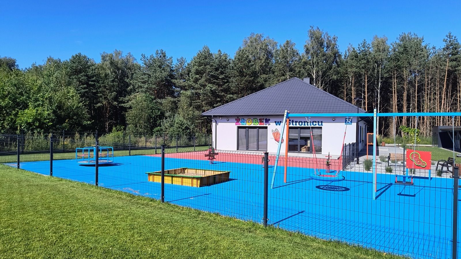 Budynek żłobka od strony trawnika, obok ogrodzony plac zabaw z niebieską nawierzchnią
